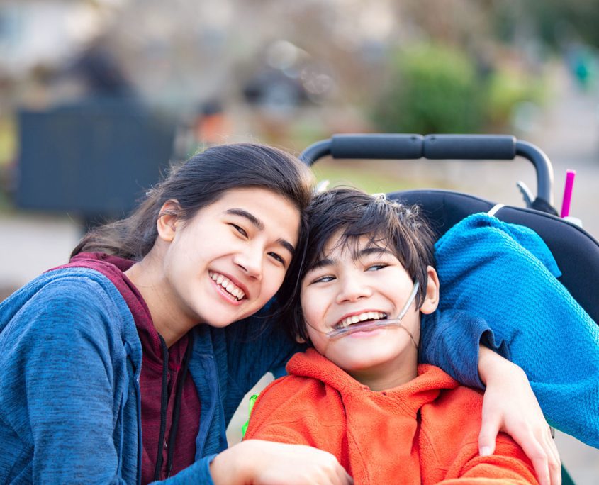 Learn More About Courage Support Services - Biracial big sister lovingly hugging disabled little brother in wheelchair outdoors